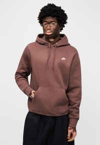 Rjava Nike jopica s sprednjim žepom, kapuco z vrvico in majhnim belim logotipom. Mehka tekstura, v kombinaciji z ohlapnimi črnimi hlačami.