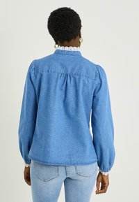 Camisa de mezclilla azul claro con un corte relajado, mangas abullonadas y un cuello con encaje. Presenta un respaldo fruncido y detalles de costuras mínimas.