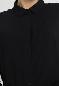 Camicia nera con colletto classico, realizzata in tessuto liscio. Presenta una pattina anteriore con bottoni scuri e senza motivi visibili.