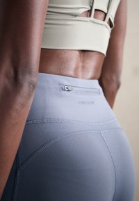 Grå atletiske leggings med en glat tekstur, der har en sikker lynlåslomme og kontrasterende syninger. Stoffet sidder højt på taljen.