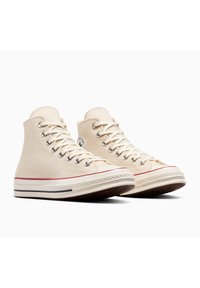 Baskets montantes en toile crème avec semelles en caoutchouc blanches. Présentent une bande rouge, un design à lacets et un patch logo sur le côté extérieur.