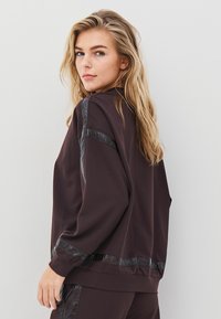 Pruun dressipluus läikivate mustade aktsentidega õlgadel ja varrukatel. Pehme kangas, mugav lõige ja ümardatud kaelus. Minimalistlik disain.