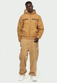 Άνδρας που φοράει καφέ ζακέτα με κουκούλα Karl Kani, μπεζ cargo παντελόνι και λευκά αθλητικά παπούτσια σε απλό ανοιχτό φόντο.
