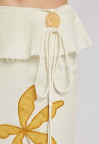 White fabric skirt with ruffled hem, featuring a yellow embroidered butterfly design, secured with a large, round yellow button and drawstring.