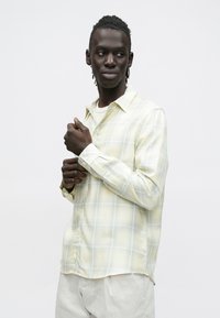 Jasnożółta koszula w kratę z długim rękawem, wykonana z miękkiej tkaniny, z kołnierzykiem i zapięciem na guziki. Gładka tekstura, luźny krój.