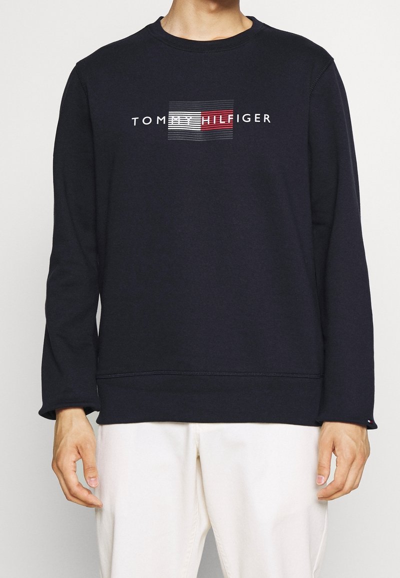 Felpa blu navy, colletto rotondo e maniche lunghe. Presenta una grafica centrale con il nome del marchio "Tommy Hilfiger" in testo bianco e rosso.