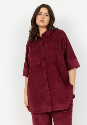 Burgundy corduroy shirt with short sleeves, button closure, two chest pockets, and a relaxed fit. Textured fabric with vertical stripes.
