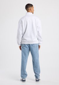 Felpa grigia con colletto alto, vestibilità rilassata e polsini a costine, abbinata a jeans azzurri chiari e sneakers. Dettagli delle cuciture visibili.