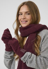 Vinröd stickad scarf och handskar gjorda av mjukt tyg, med ribbat mönster och en läderdetalj på handskarna. Grå tröja under.