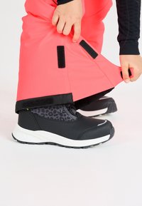 Schwarze wasserdichte Stiefel mit einem strukturierten grauen Leopardenmuster im Innenbereich, kombiniert mit leuchtend pinken Latzhosen, die von einer Hand angepasst werden.