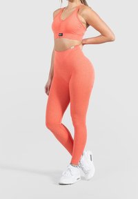 Dvoudílný sportovní outfit v živé korálové barvě. Zkrácený top má výstřih do V a silné ramínka; legíny s vysokým pasem jsou jemně žebrované pro texturu.