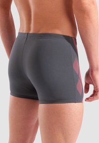 Mörkgrå badshorts i slät tyg, med ett rött diamantmönster på sidan och en snug passform med en ren midja.