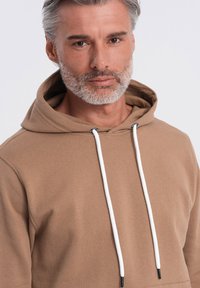 Felpa marrone realizzata in tessuto morbido e liscio, con tasca a marsupio. Presenta cordini bianchi con dettagli neri e una vestibilità comoda.