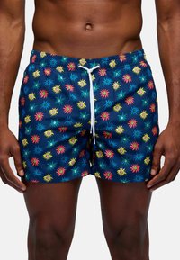 Un uomo sta in piedi indossando costumi da bagno colorati con un motivo a raggio di sole in rosa, giallo e blu su uno sfondo scuro.