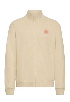 Veste polaire beige à fermeture éclair intégrale avec poignets et ourlet côtelés, présentant un petit dessin de soleil orange et le texte "SUN SEEKER" sur le côté gauche de la poitrine.