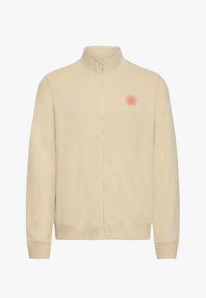 Veste polaire beige à fermeture éclair intégrale avec poignets et ourlet côtelés, présentant un petit dessin de soleil orange et le texte "SUN SEEKER" sur le côté gauche de la poitrine.