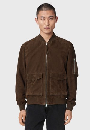 Männliches Modell trägt eine dunkelbraune Wildleder-Bomberjacke mit Fronttaschen und Reißverschluss, kombiniert mit schwarzen Hosen, stehend vor einem schlichten Hintergrund.