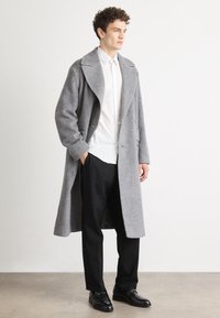 Grå hørvoks lang frakke med stor krave, båret over en hvid skjorte og sorte bukser. Sorte sko fuldender outfittet.