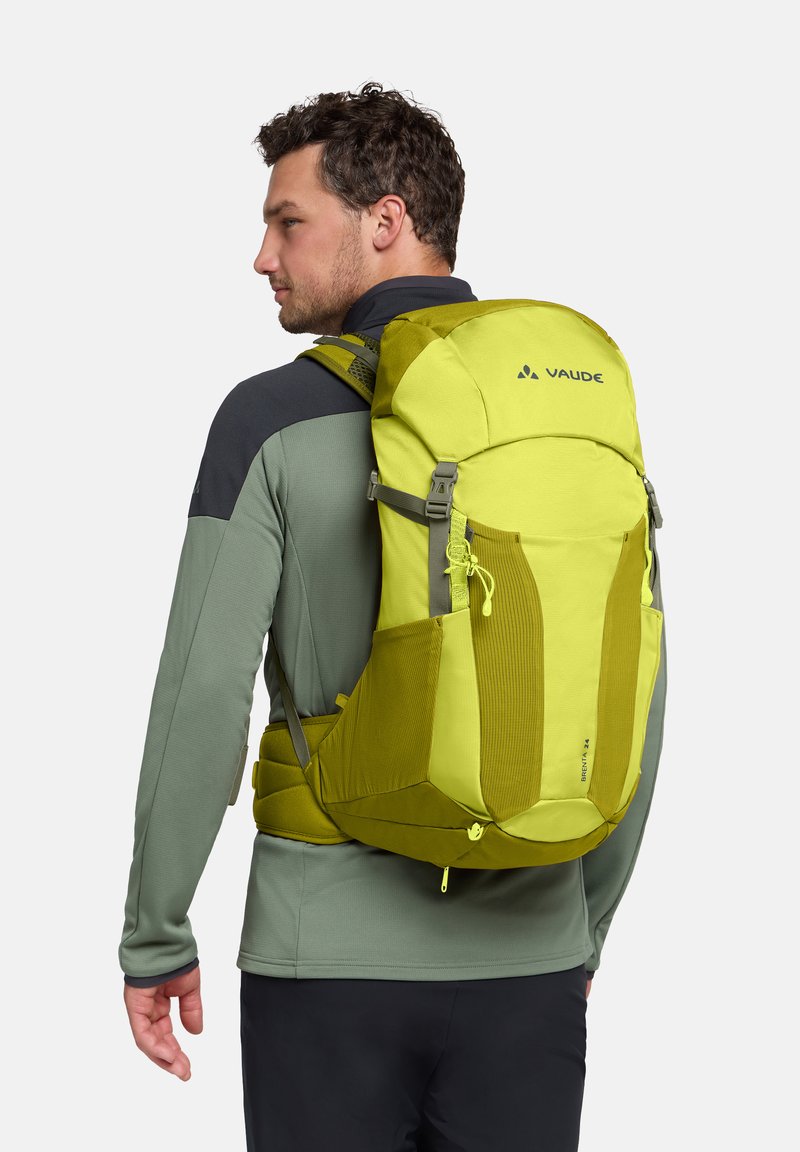 Homme portant un sac à dos de randonnée vert citron avec bretelles rembourrées et ceinture de hanche, habillé d'une veste à manches longues verte et noire et d'un pantalon noir.