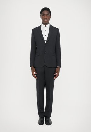 Mørkegrå jakkesæt med en skræddersyet jakke, matchende bukser og en hvid skjorte med sorte knapper. Sorte dressko fuldender outfittet.