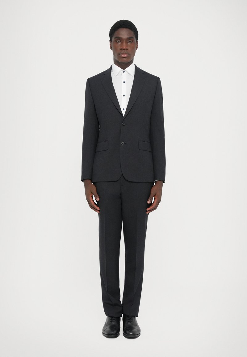 Mørkegrå jakkesæt med en skræddersyet jakke, matchende bukser og en hvid skjorte med sorte knapper. Sorte dressko fuldender outfittet.