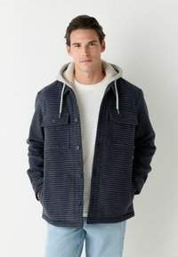 Marineblau karierter Jacke mit grauem Kapuze, ausgestattet mit einem weichen, strukturierten Innenfutter, zwei Brusttaschen und Knopfverschlüssen, getragen über einem hellen Hemd.