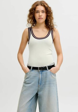 Fiatal nő göndör hajjal, fehér ujjatlan felsőt visel fekete szegéllyel és laza világoskék farmert fekete övvel, kezét zsebében tartja.