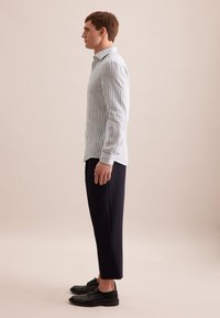 Gestreepte blauwe en witte blouse met lange mouwen, gecombineerd met korte donkere broek en zwarte schoenen, zijaanzicht tegen een neutrale achtergrond.
