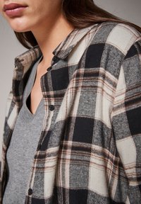 Camicia di flanella a quadri neri e crema con accenti marroni, caratterizzata da colletto, chiusura con bottoni e indossata sopra un top grigio a scollo a V.