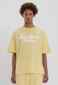 T-shirt en coton jaune à manches courtes et col rond, avec un texte en script blanc "Low Lights Studios" sur le devant. Texture douce.
