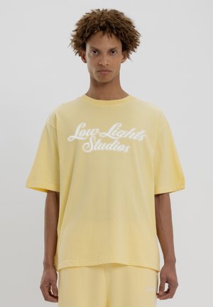 T-shirt en coton jaune à manches courtes et col rond, avec un texte en script blanc "Low Lights Studios" sur le devant. Texture douce.