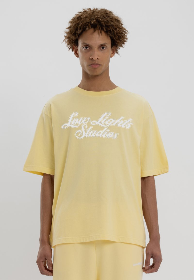T-shirt en coton jaune à manches courtes et col rond, avec un texte en script blanc "Low Lights Studios" sur le devant. Texture douce.