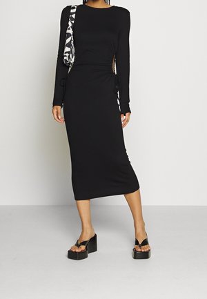 Robe noire à manches longues avec ruching sur les côtés et cordons de serrage, longueur midi. Portée avec des sandales à plateforme noires et un sac à épaule à motifs.