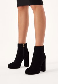 Botas de ante negras hasta el tobillo con puntera cuadrada, tacón alto en bloque y detalle de cremallera lateral. Textura suave con un diseño minimalista.