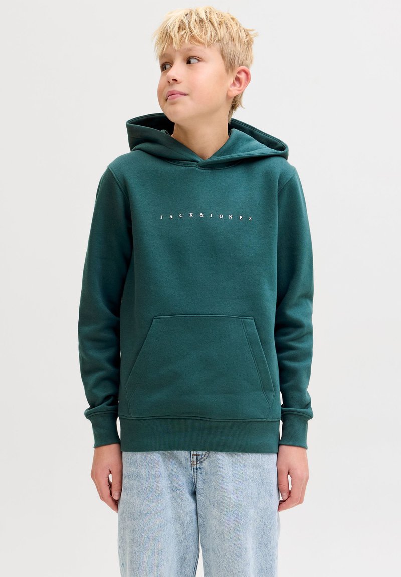Ragazzo biondo che indossa una felpa con cappuccio Jack & Jones verde scuro e jeans azzurri, guarda leggermente di lato su uno sfondo semplice.