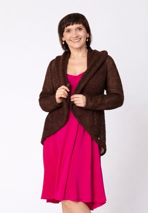 Kobieta uśmiechająca się, ubrana w jasnoróżową sukienkę i brązowy dzianinowy kardigan, stojąca na tle jednolitego białego tła.