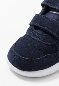 Marinblå mockaskor med kardborreband, perforerat tåparti, texturerad överdel och vit gummisula. Närbild som fokuserar på skodetaljer.