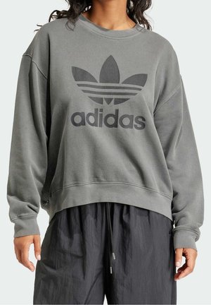Osoba ubrana w luźną szarą bluzę Adidas z logo trefoil i czarne spodnie z troczkami, widoczna od szyi do połowy ud.