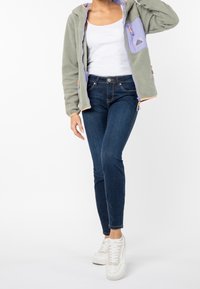Chaqueta de forro polar con cremallera en verde claro con detalles en lavanda y bolsillos, combinada con unos vaqueros azul oscuro y zapatillas blancas.