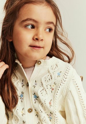 Young girl with long brown hair wearing a cream knitted cardigan with floral embroidery, looking to the side against a plain background.