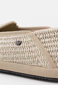 Beige vävd tyg slip-on sko med en svart metallaccent och en texturerad sula. Visar en rundad design och förstärkt tygkanter.