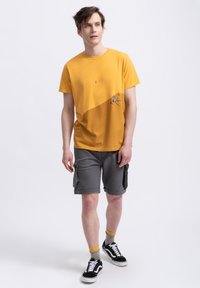 Geel katoen T-shirt met een grafisch ontwerp op de linksonder; gecombineerd met grijze cargoshorts en zwarte sneakers met witte zolen.