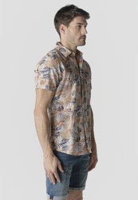 Hombre con camisa de manga corta abotonada con patrones de hojas en naranja y azul, y pantalones cortos de mezclilla remangados, de pie de perfil.