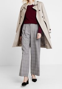 Beige trenchcoat met zwarte knopen over een bordeauxrode trui en een witte blouse. Grijs geruite hooglopende wijde broek en zwarte schoenen.