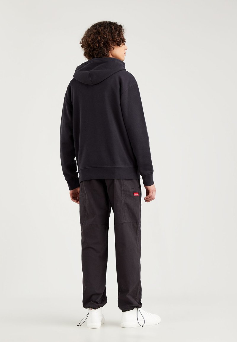 Sudadera oscura con bolsillo tipo canguro, combinada con pantalones cargo oscuros que tienen una pequeña etiqueta roja. Zapatillas blancas añaden contraste.