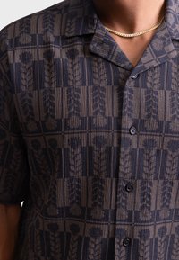 Camicia a maniche corte in navy e beige, con un motivo botanico testurizzato e bottoni scuri. Design con colletto e vestibilità rilassata.