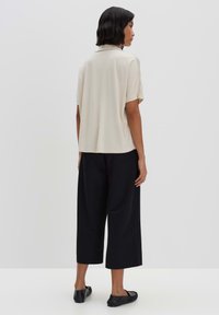 Beige Bluse mit kurzen Ärmeln, lockerem Schnitt und Kragen, kombiniert mit schwarzen weiten Hosen und flachen Schuhen, von hinten betrachtet.