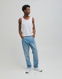 Nuori mies seisoo kädet taskuissa, päällään valkoinen hihaton toppi, vaaleansiniset farkut ja valkoiset tennarit yksinkertaista taustaa vasten.