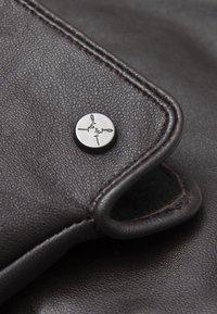 Tamni kožni detalj s zaobljenim rubom, vidljivim šavovima i metalnom tipkom s logotipom. Tekstura je glatka s blagom zrnatošću.