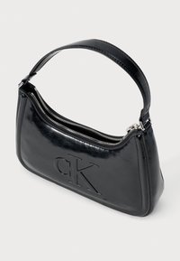 Čierna kožená ramenná taška s zakriveným tvarom, embosované logo "CK", strieborný zips a hladký, lesklý povrch. Rukoväť je umiestnená na vrchu.